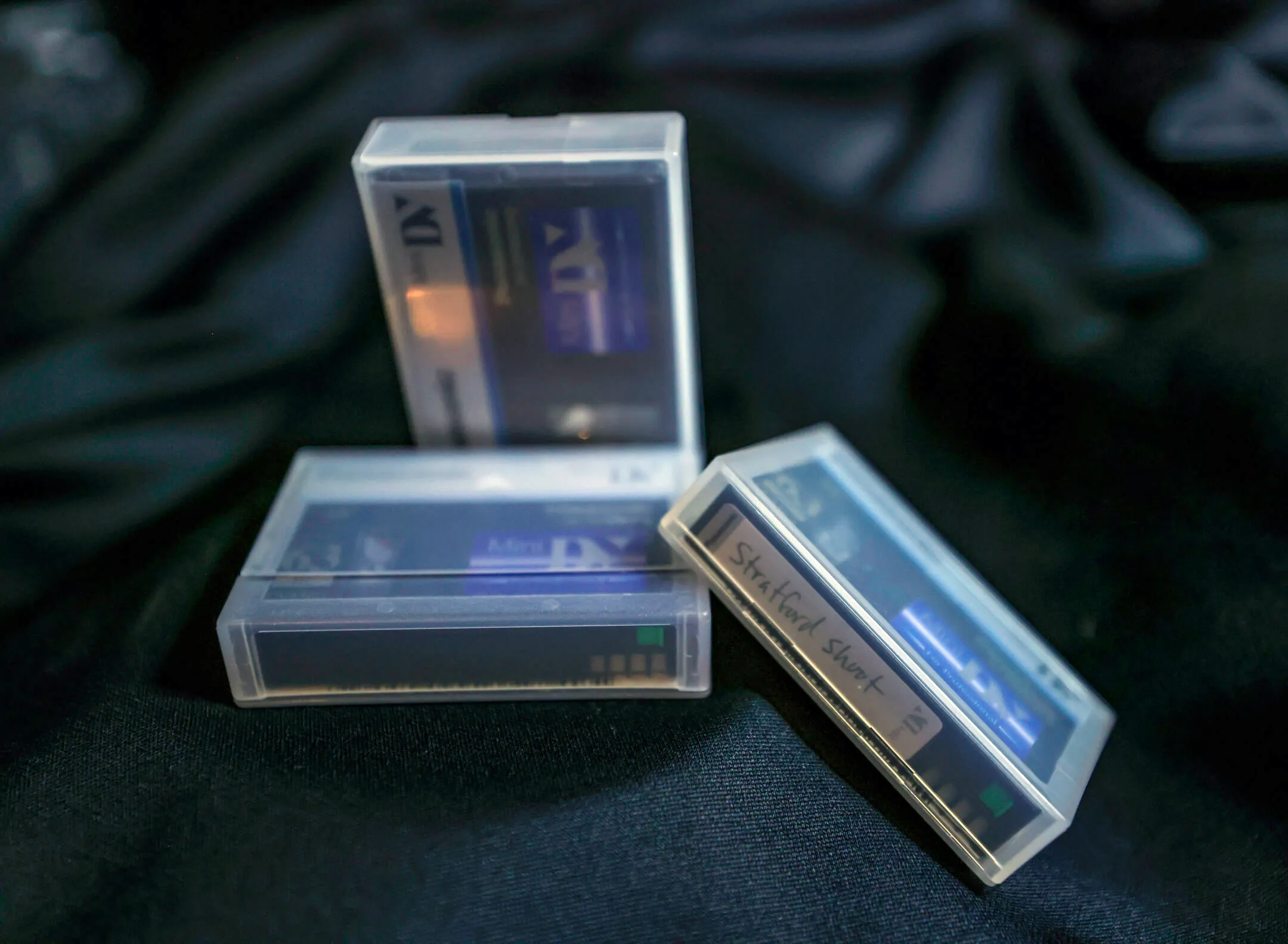 Three MiniDV tapes in plastic cases on a dark cloth background, one labeled by hand 'Stratford Short'.