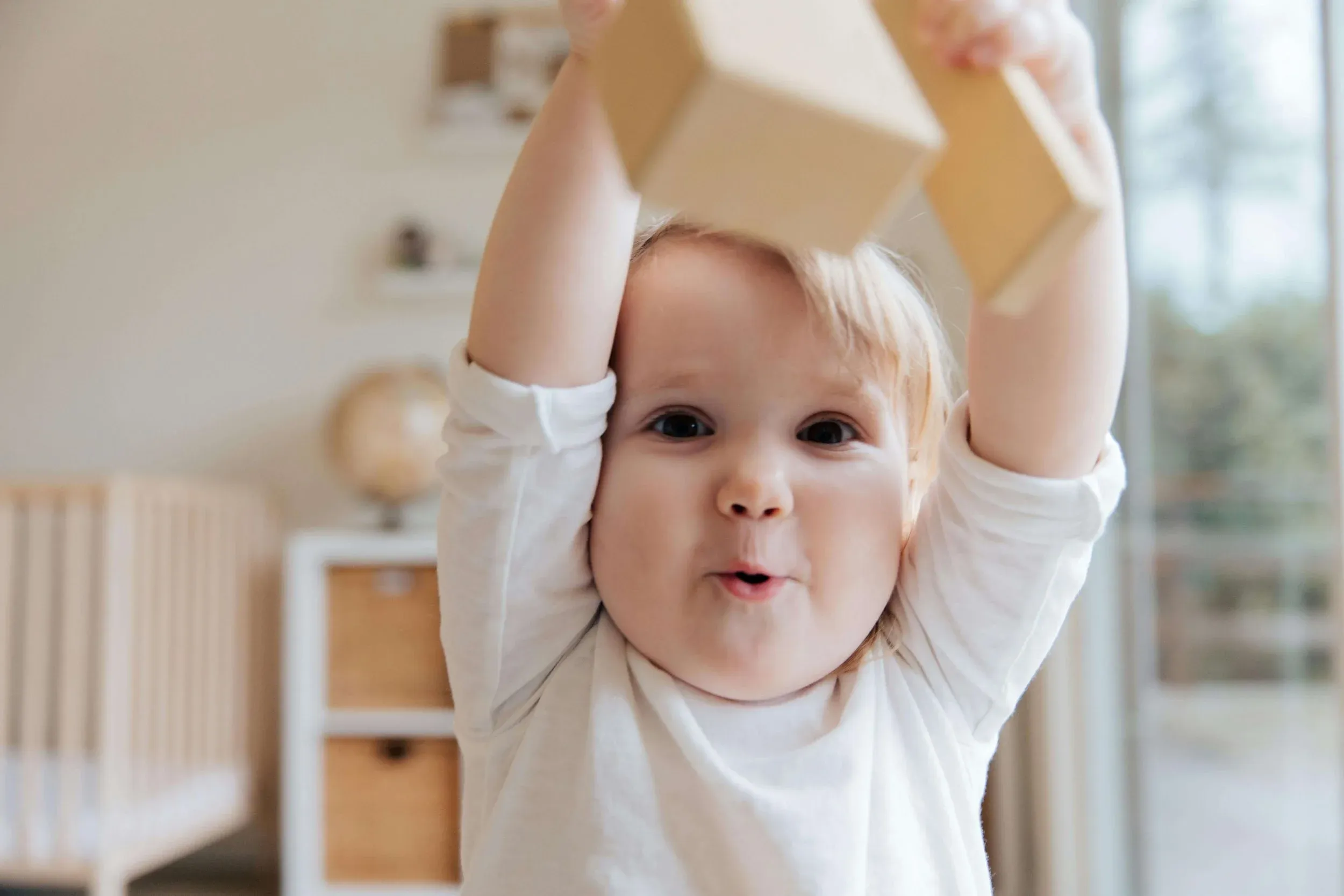 A toddler with blonde hair holds two wooden blocks overhead, mouth open mid-motion, captured just before the next moment lands.