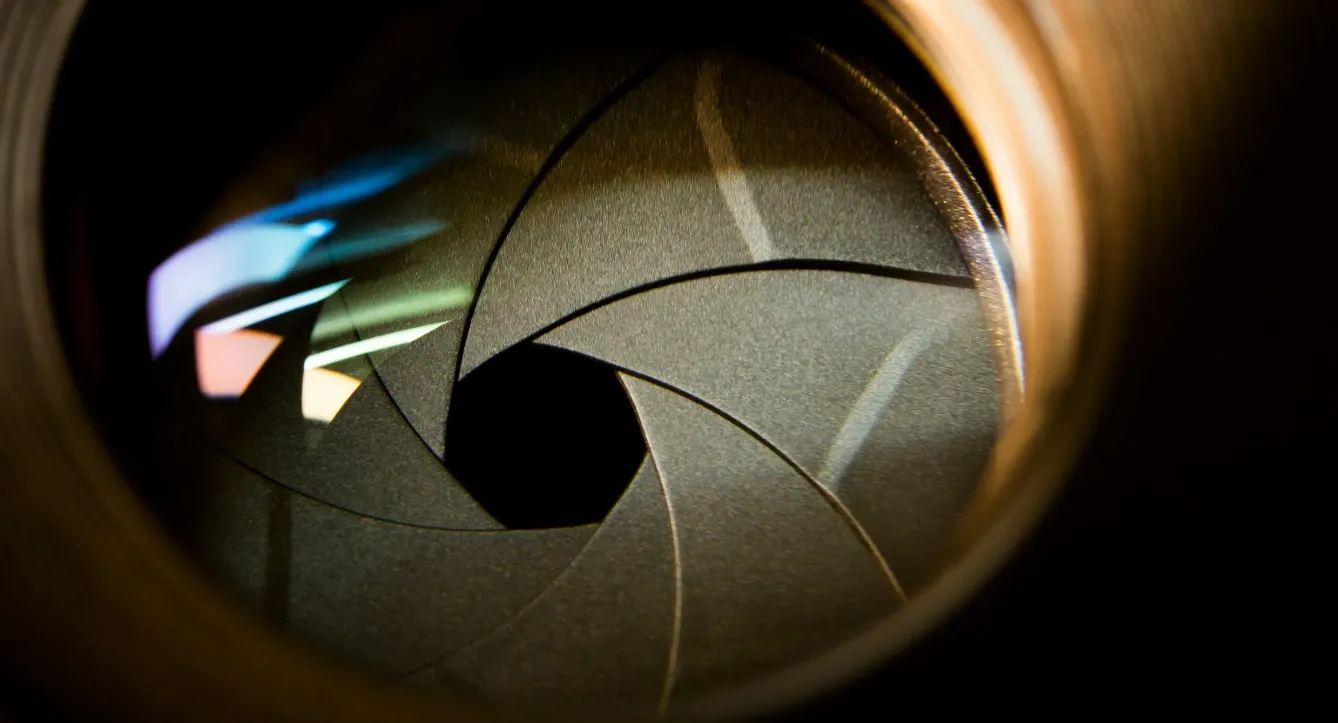 Extreme close-up looking through a camera lens at the aperture diaphragm blades forming a spiral opening, with colorful light reflections.