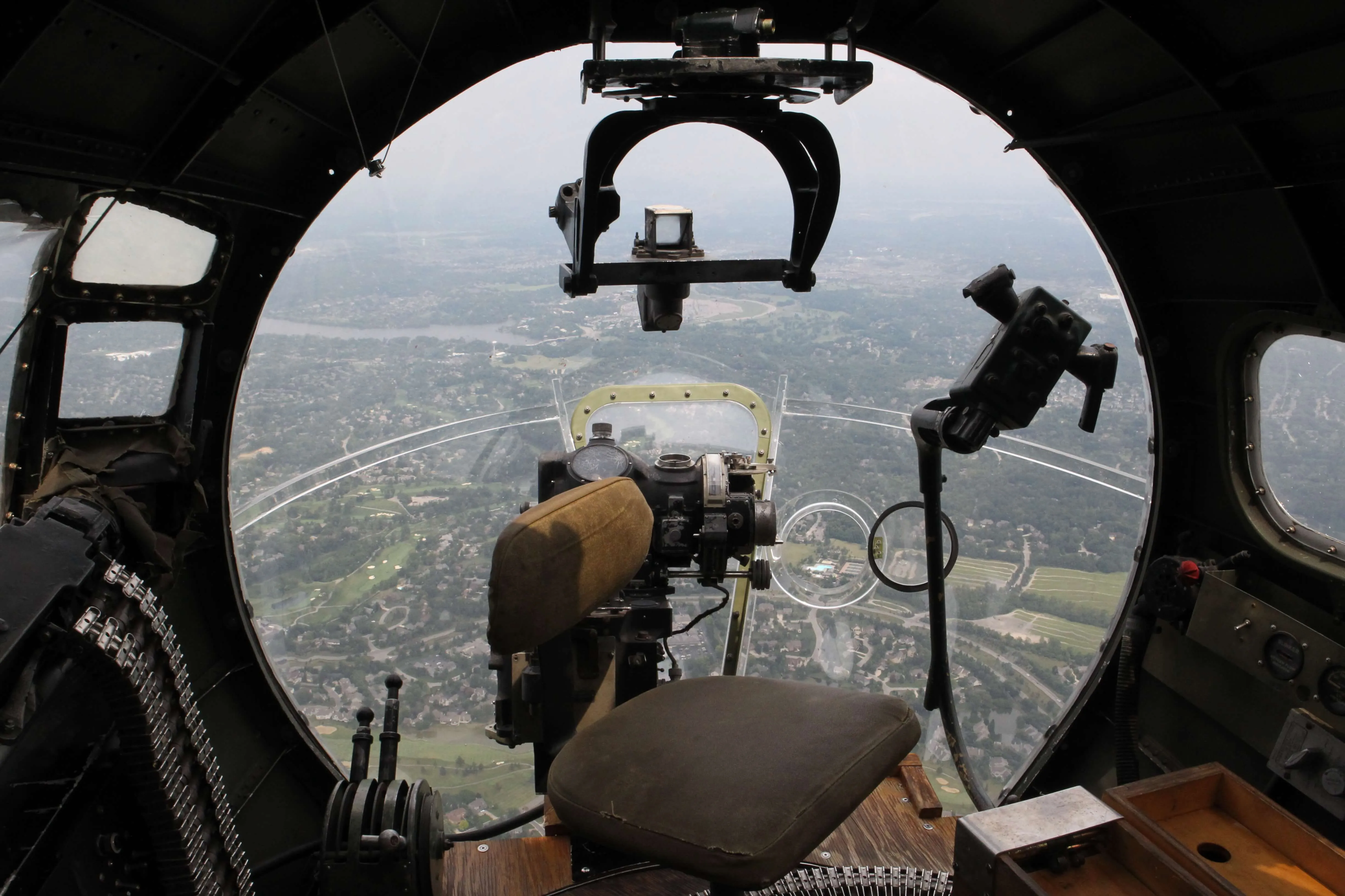 View from inside the nose turret of a WWII bomber, looking out over a hazy aerial landscape through the gunner's sighting equipment.