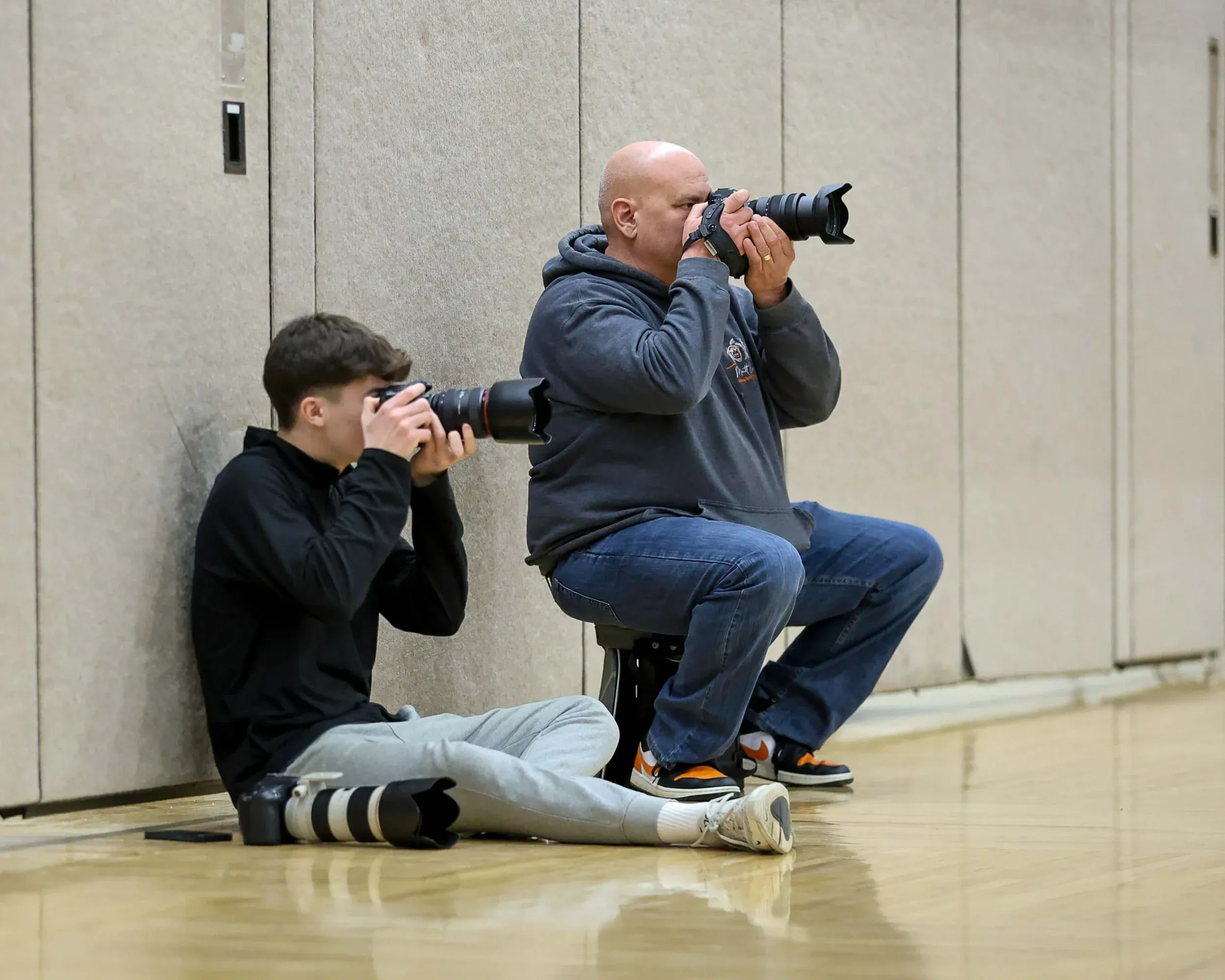 A bald man in a gray hoodie and a teenage boy in a black hoodie sit low against a gym wall, both aiming large telephoto lenses to photograph a basketball game from floor level.