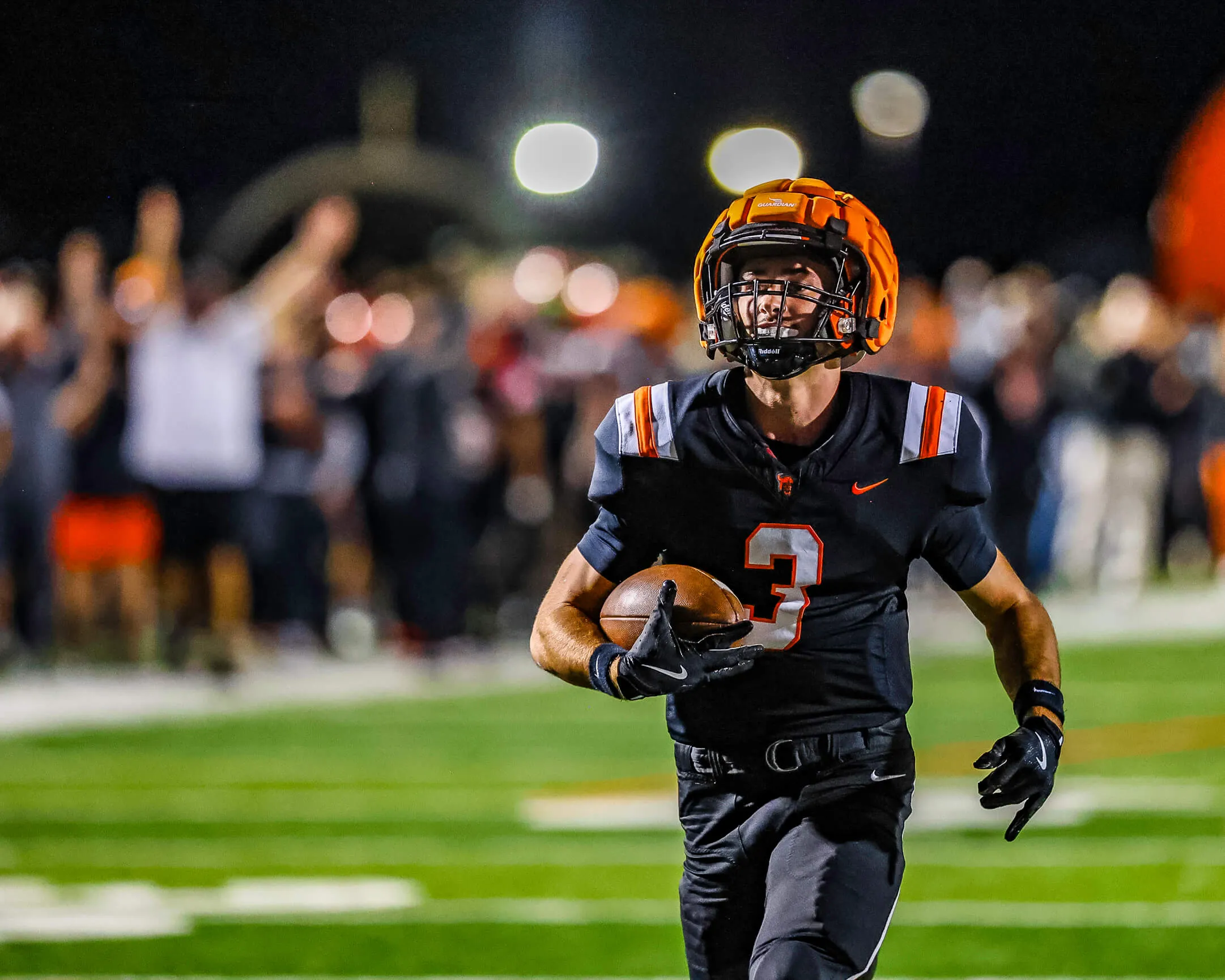 A high school football player in an orange helmet running with the ball under stadium lights at night, sharply frozen against a blurred, cheering crowd.