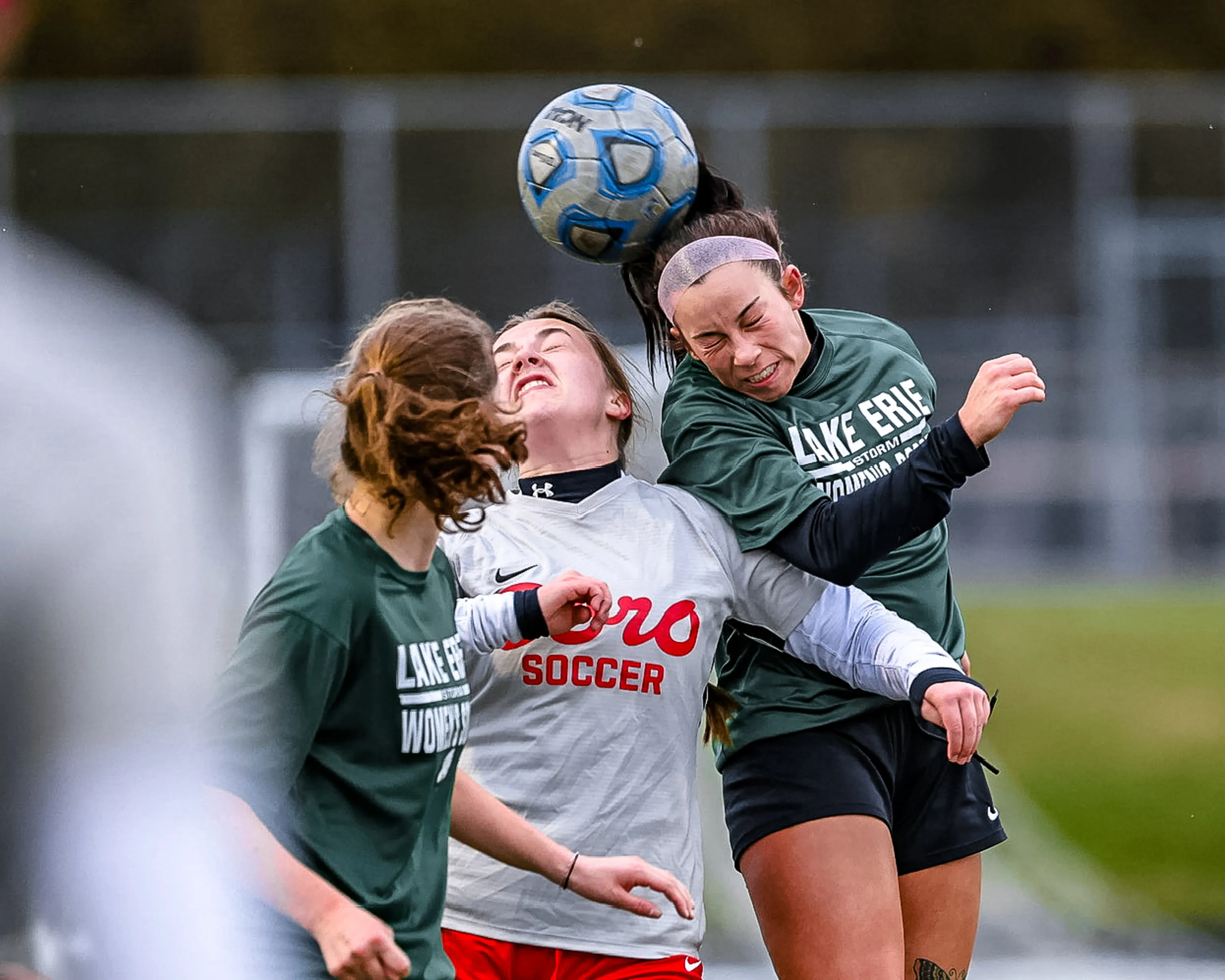 Three college soccer players colliding in a header duel over the ball, all sharply frozen mid-air with a blurred stadium background.