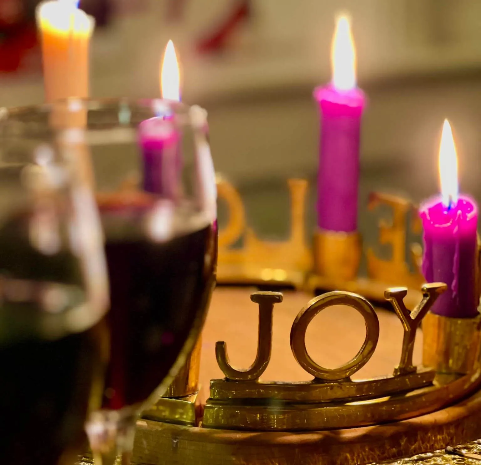 Close-up of a glass of red wine beside a lit Advent wreath with purple candles and a brass 'JOY' ornament, shot in warm, low indoor light.