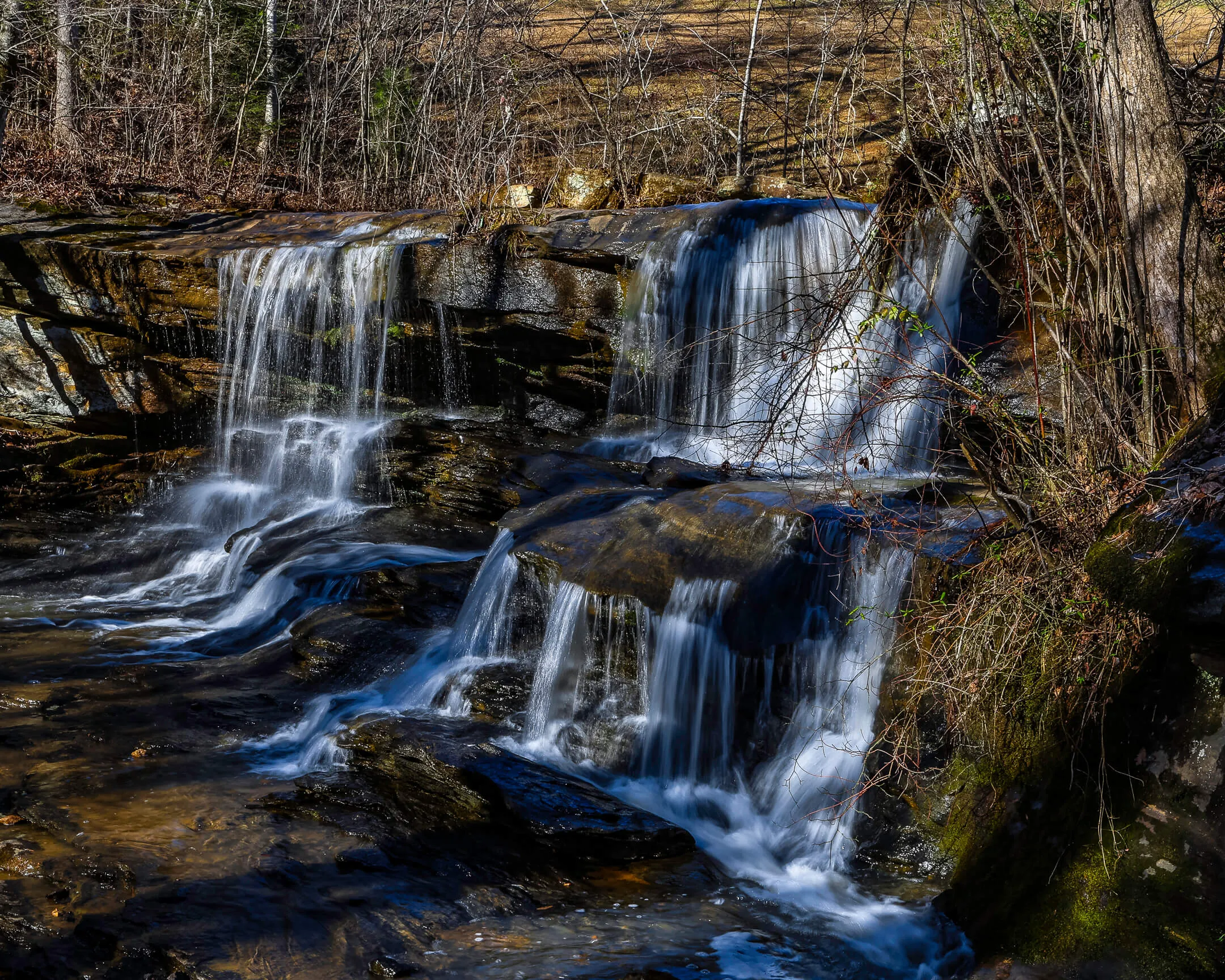 A long-exposure waterfall over layered rock ledges in a wooded creek, water rendered smooth and silky.