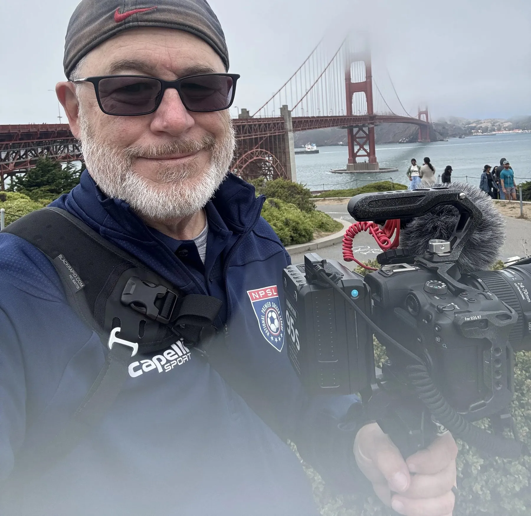 TMac smiling at the camera with the Golden Gate Bridge in the fog behind him, a camera and shotgun mic rig hanging around his neck.