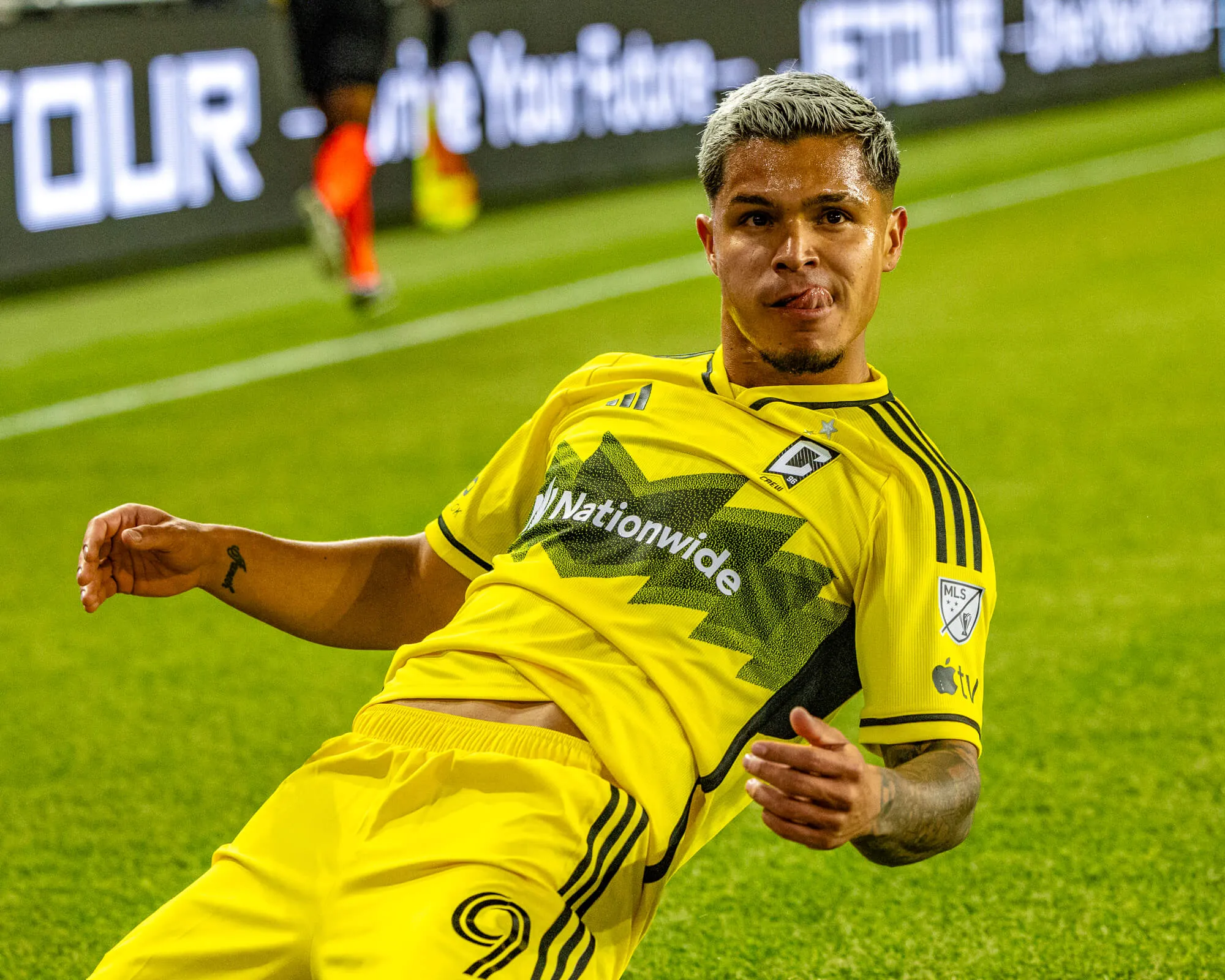A Columbus Crew MLS player in a yellow kit celebrating on the pitch, tongue out, arm extended, with blurred LED perimeter boards behind him.