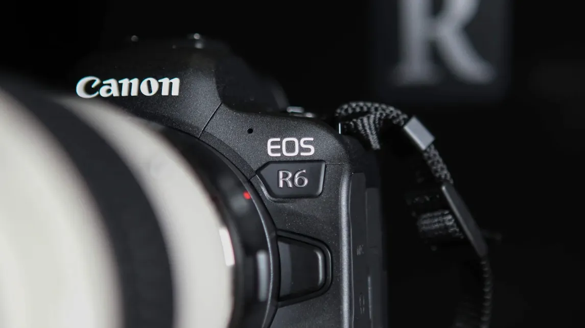 A close-up of a Canon EOS R6 camera body with a white telephoto lens, dramatically lit against a black background.