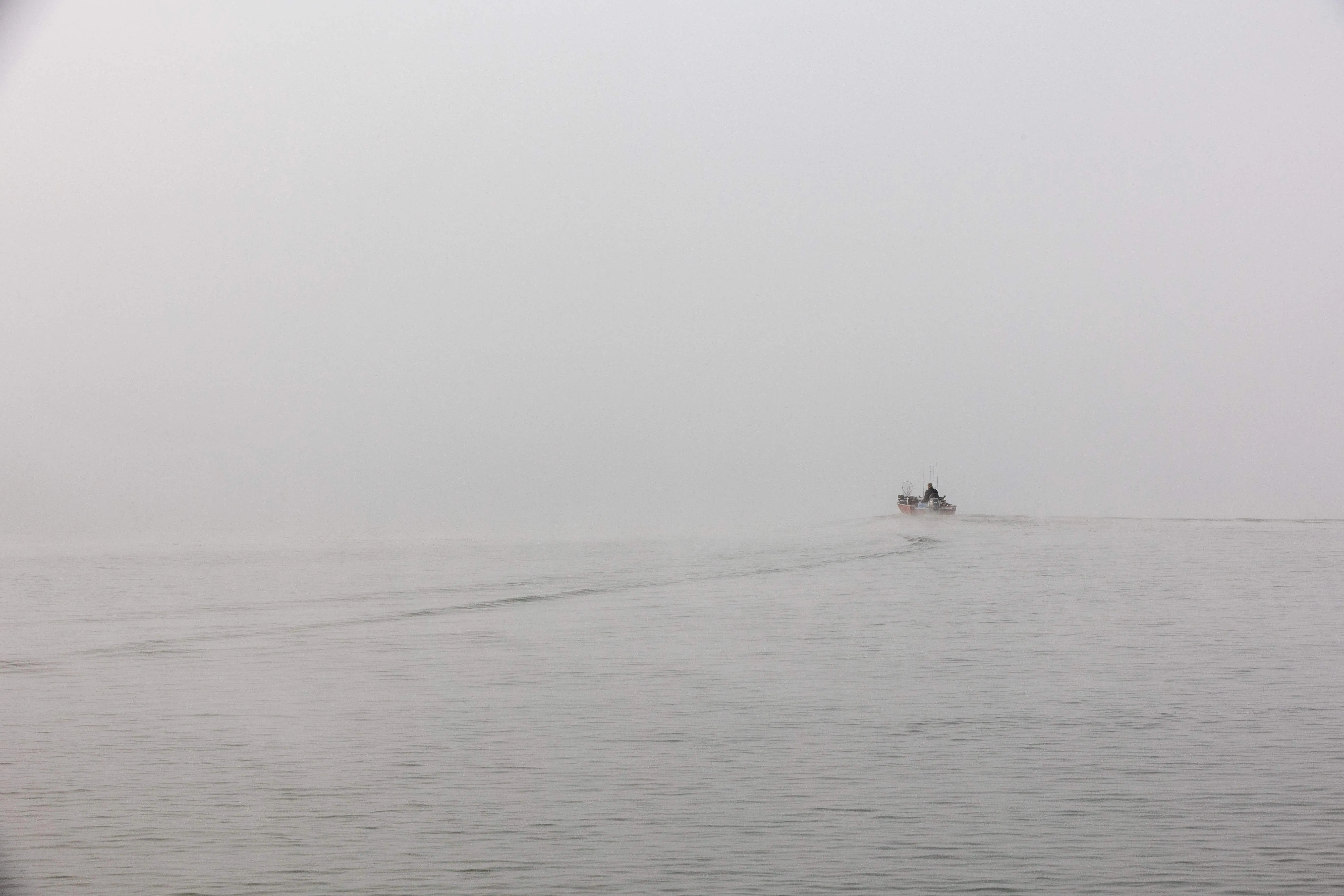 A small fishing boat with an angler aboard, cutting a wake through calm water inside dense gray fog.