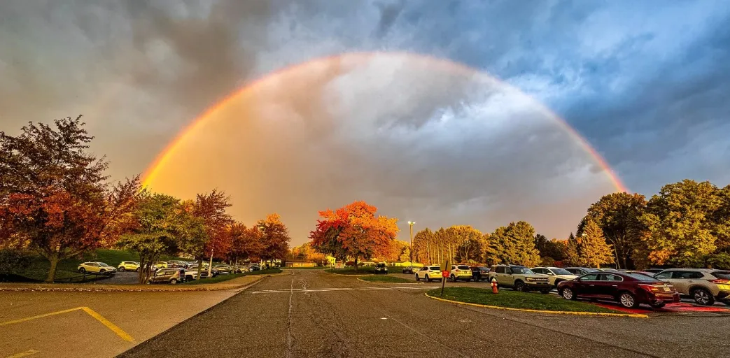 A full rainbow arcing over a school parking lot at sunset, fall foliage on the trees around the lot.