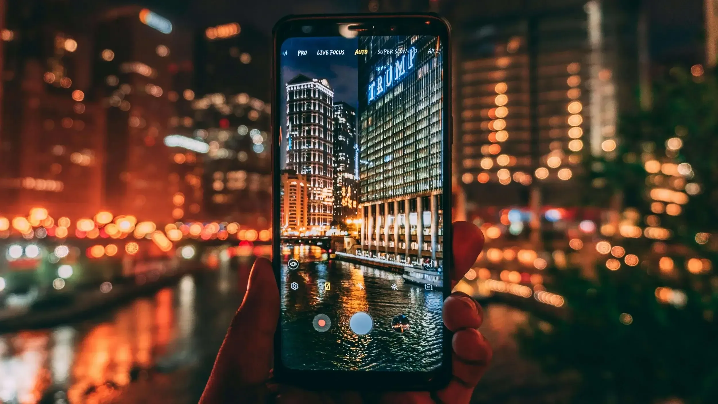 A hand holding a smartphone, camera app open, framing a nighttime Chicago cityscape with illuminated skyscrapers reflecting off the river.