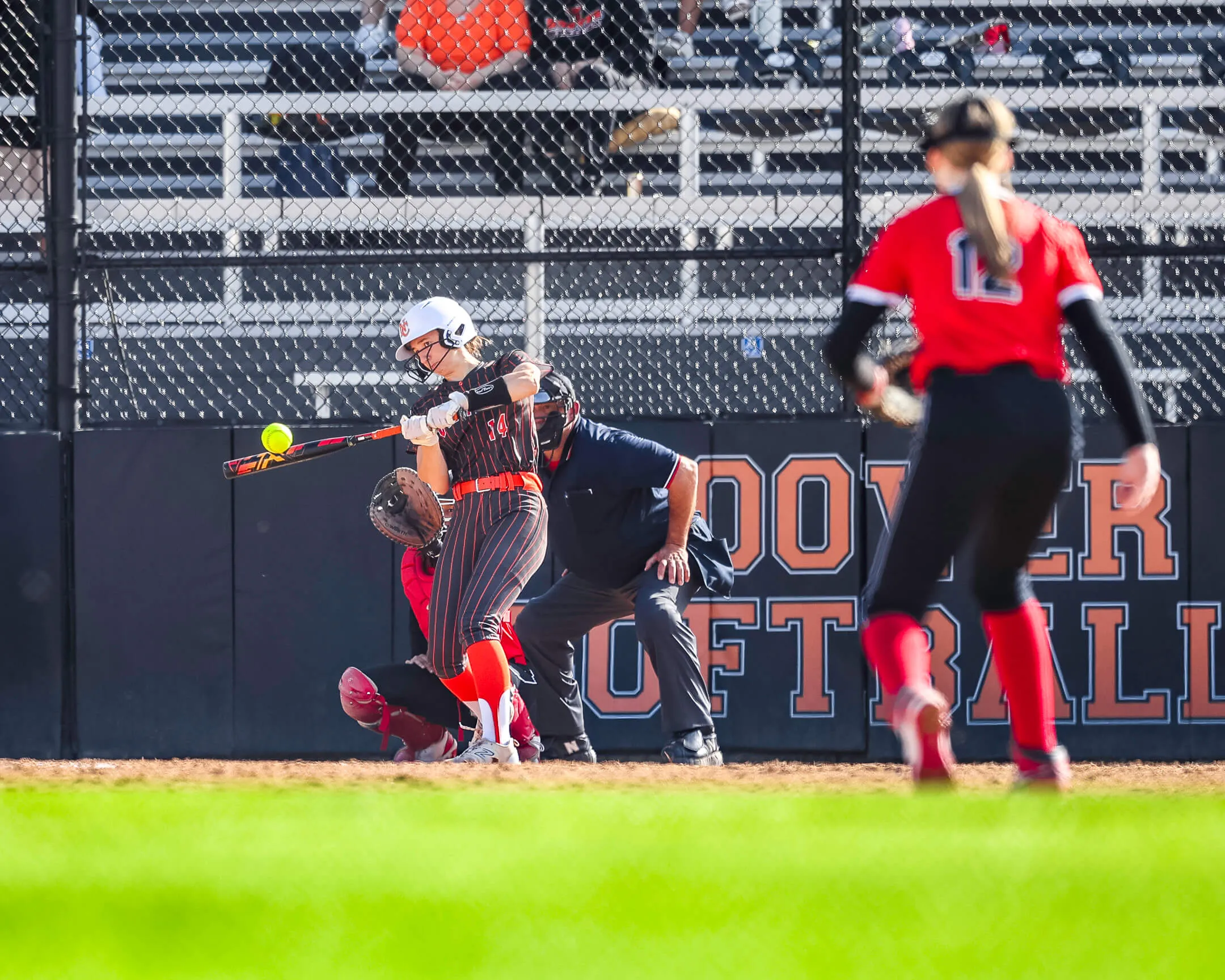 A softball batter connecting mid-swing, catcher and umpire crouched behind home plate, shot from a low belly-level angle with blurred grass in the foreground.