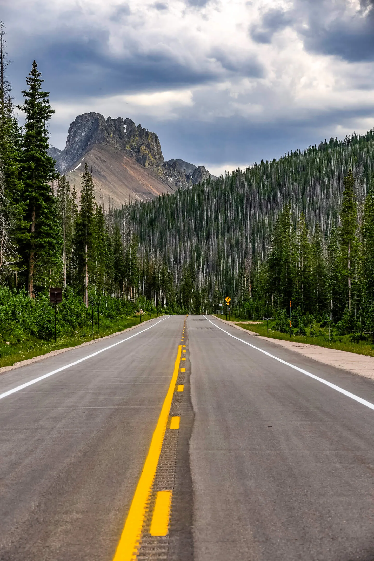 A two-lane mountain road leading toward a jagged rock peak, flanked by evergreen forest under a cloudy sky.