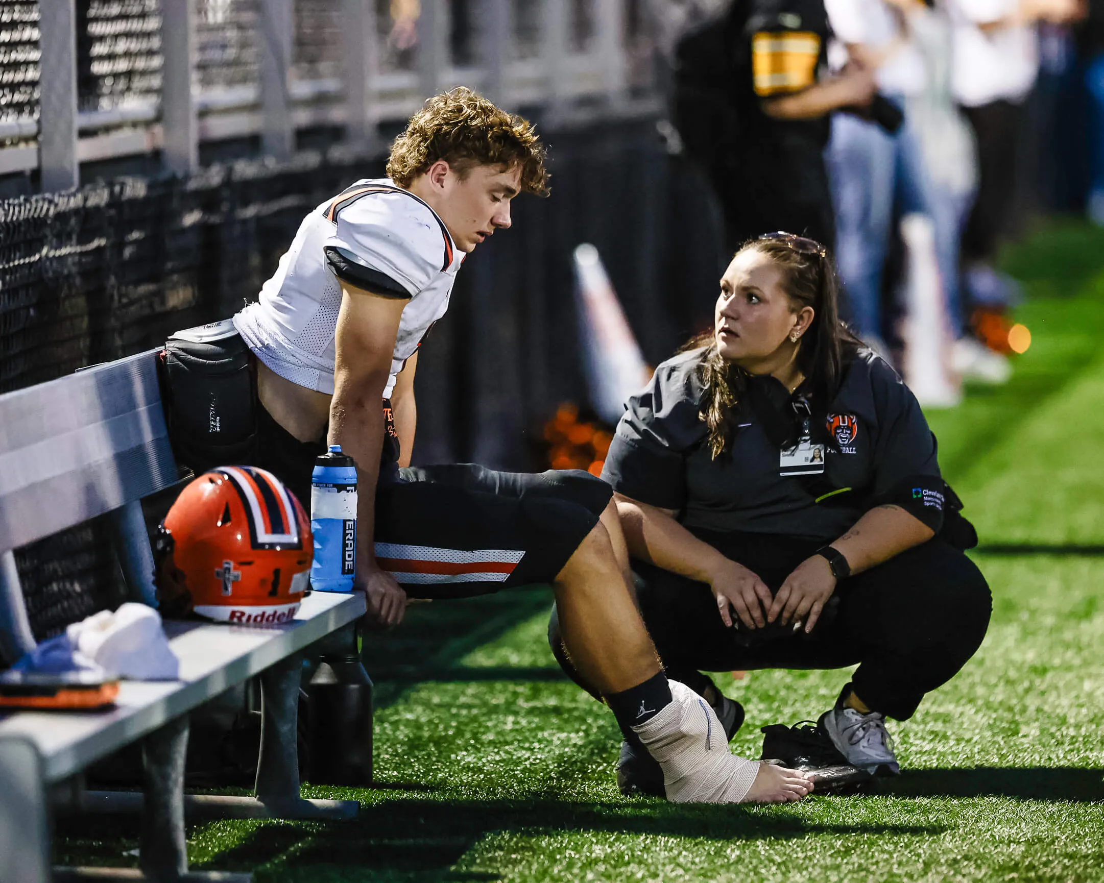 An injured football player sitting on the sideline bench, ankle wrapped, talking with an athletic trainer crouched in front of him.
