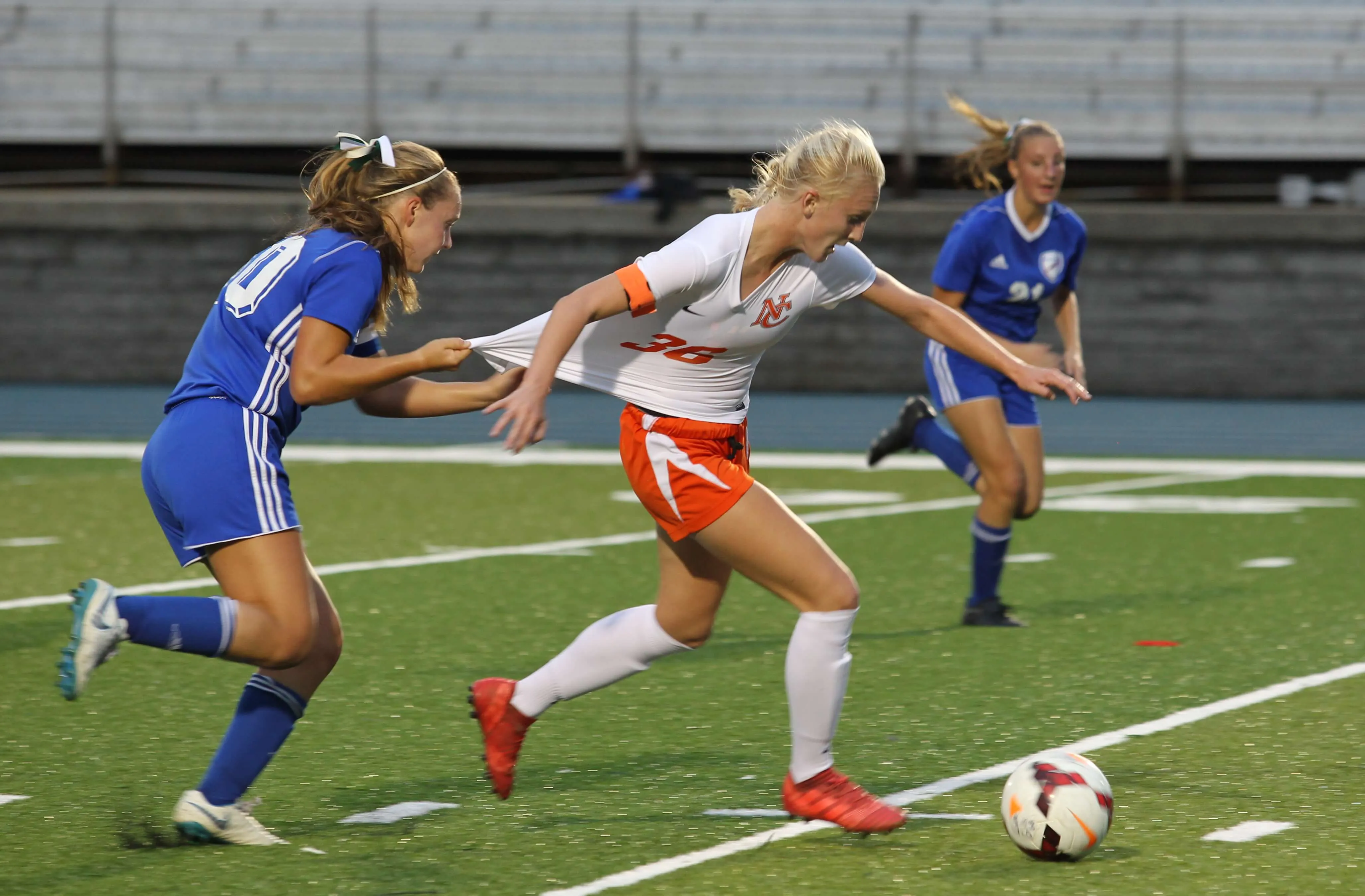 A girl in a white and orange soccer jersey, number 36, breaking away with the ball as a defender in blue grabs and pulls her jersey from behind, motion blur visible along the edges of both players.
