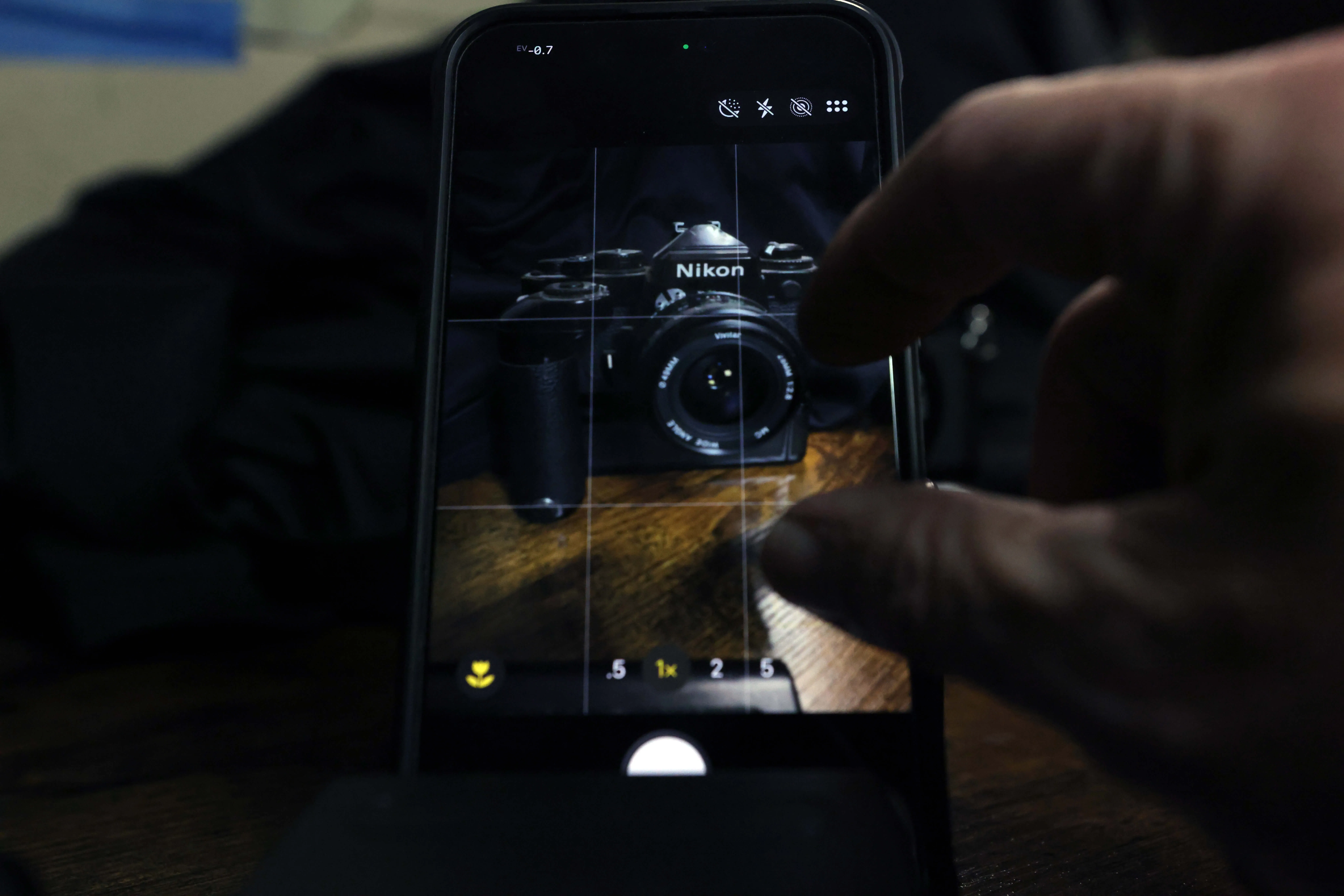 A phone screen framing a vintage Nikon film camera with a 50mm lens through the Camera app's grid, a hand's fingers reaching in to pinch-zoom.