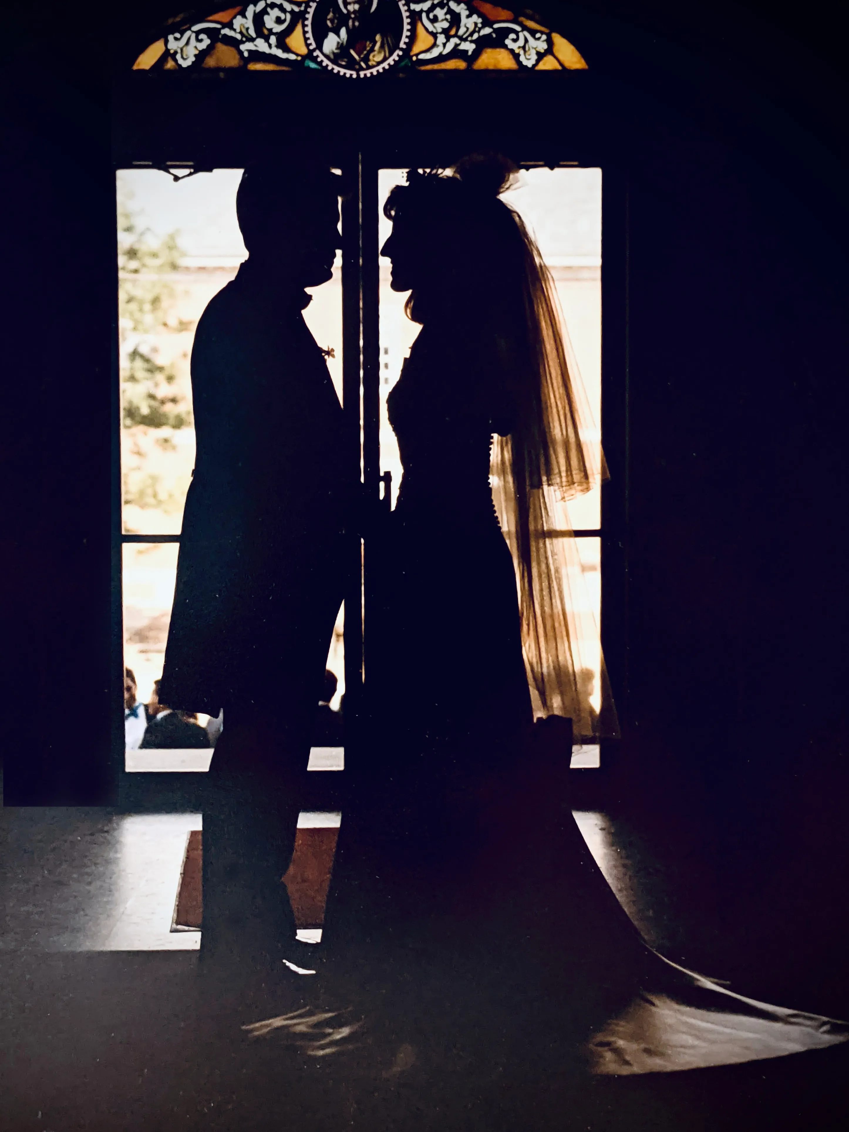 A silhouetted bride and groom facing each other in a church doorway, backlit by bright daylight streaming through the open doors.